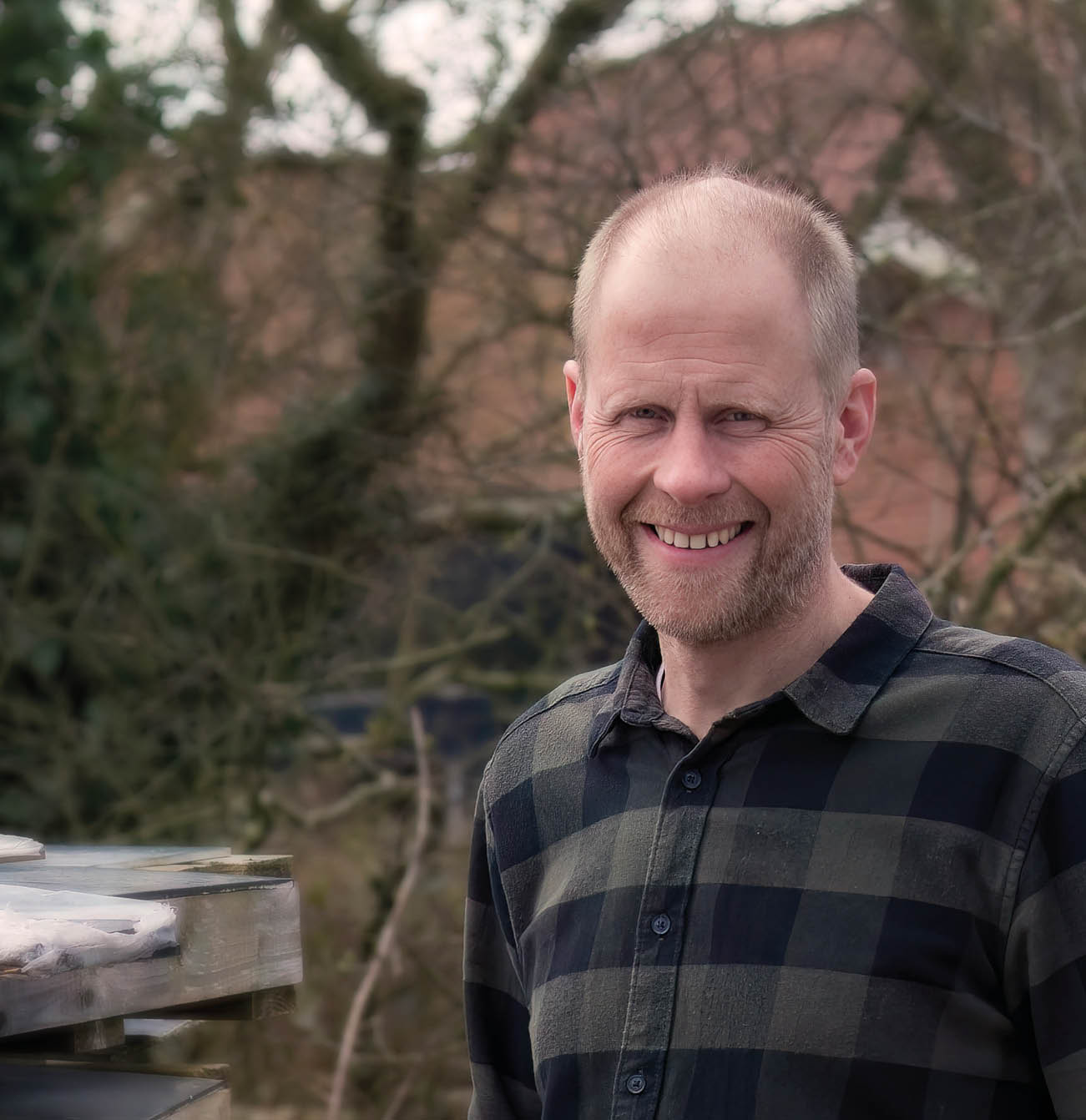 A smiling person with short hair wearing a checkered shirt stands outdoors near a wooden structure with trees and a building in the background. Beskrivelse genereret af AI