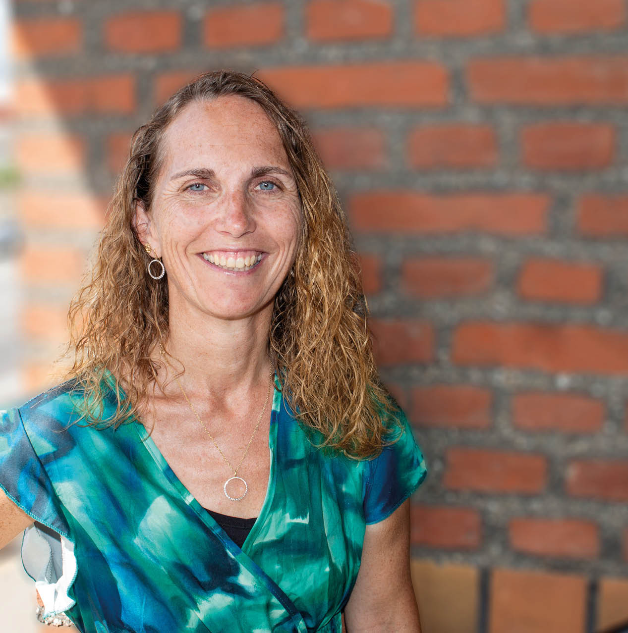 A person with curly shoulder-length hair wearing a blue and green patterned top is smiling in front of a brick wall. Beskrivelse genereret af AI