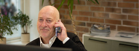 A senior with a bald head is smiling and talking on a mobile phone in a cozy indoor setting with plants and stacked magazines in the background. Beskrivelse genereret af AI