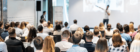 A person is giving a presentation to a seated audience in a conference room with a large screen displaying images. Beskrivelse genereret af AI
