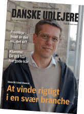 A person wearing glasses, a light sweater, and a dark jacket stands indoors near a brick wall and window, featured on the cover of a Danish magazine titled “Danske Udlejere" with text about succeeding in a difficult industry. Beskrivelse genereret af AI